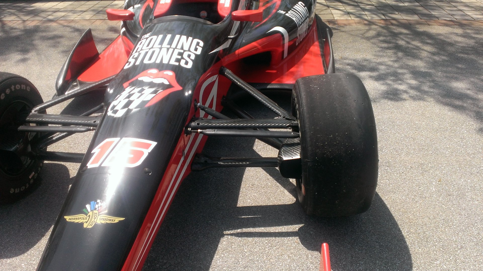 A Dallara DW12 IndyCar front corner, where the two wishbones are visible as bare rods running from the tub to the upright. Nothing here has to clear an engine, a driveshaft or a crash structure, which is why racing cars take the expensive layout almost without exception.