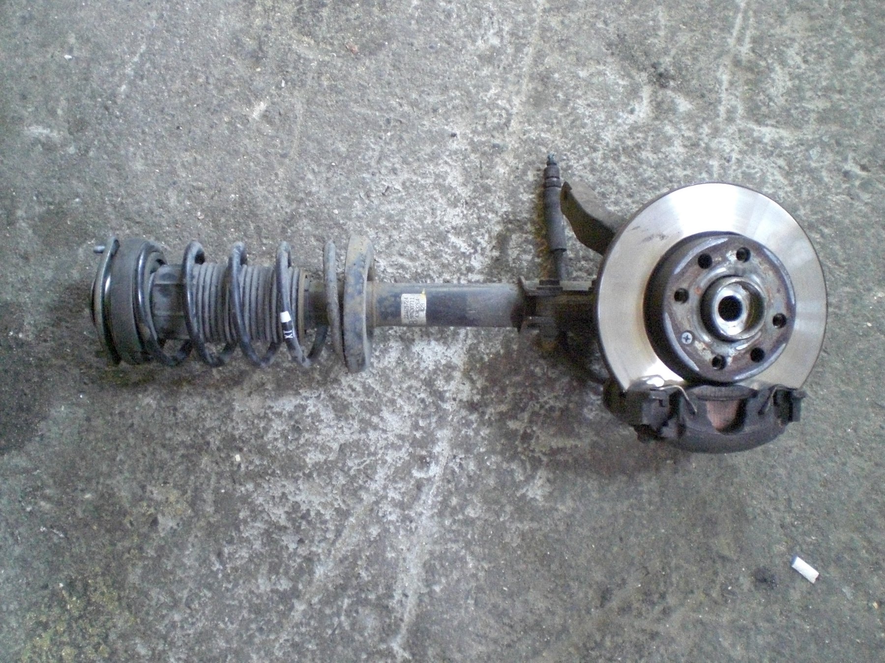 The other layout, out of the car: a MacPherson strut assembly with the spring, damper, steering knuckle, hub and disc all hanging off one unit. There is no upper arm because the strut does that job itself, which is exactly why this design is cheap to build and quick to fit on a production line.