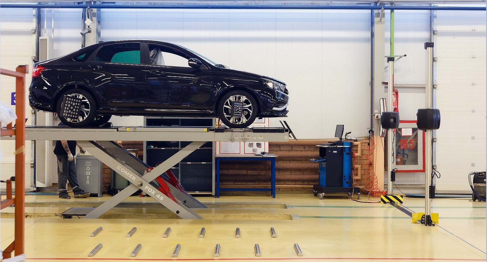 A four-wheel alignment in progress: reflective targets on all four wheels, camera towers at the end of the bay, turn plates under the fronts. The rear targets are the point — without reading the rear axle you cannot find the thrust line, and without the thrust line the front numbers are aimed at the wrong reference.